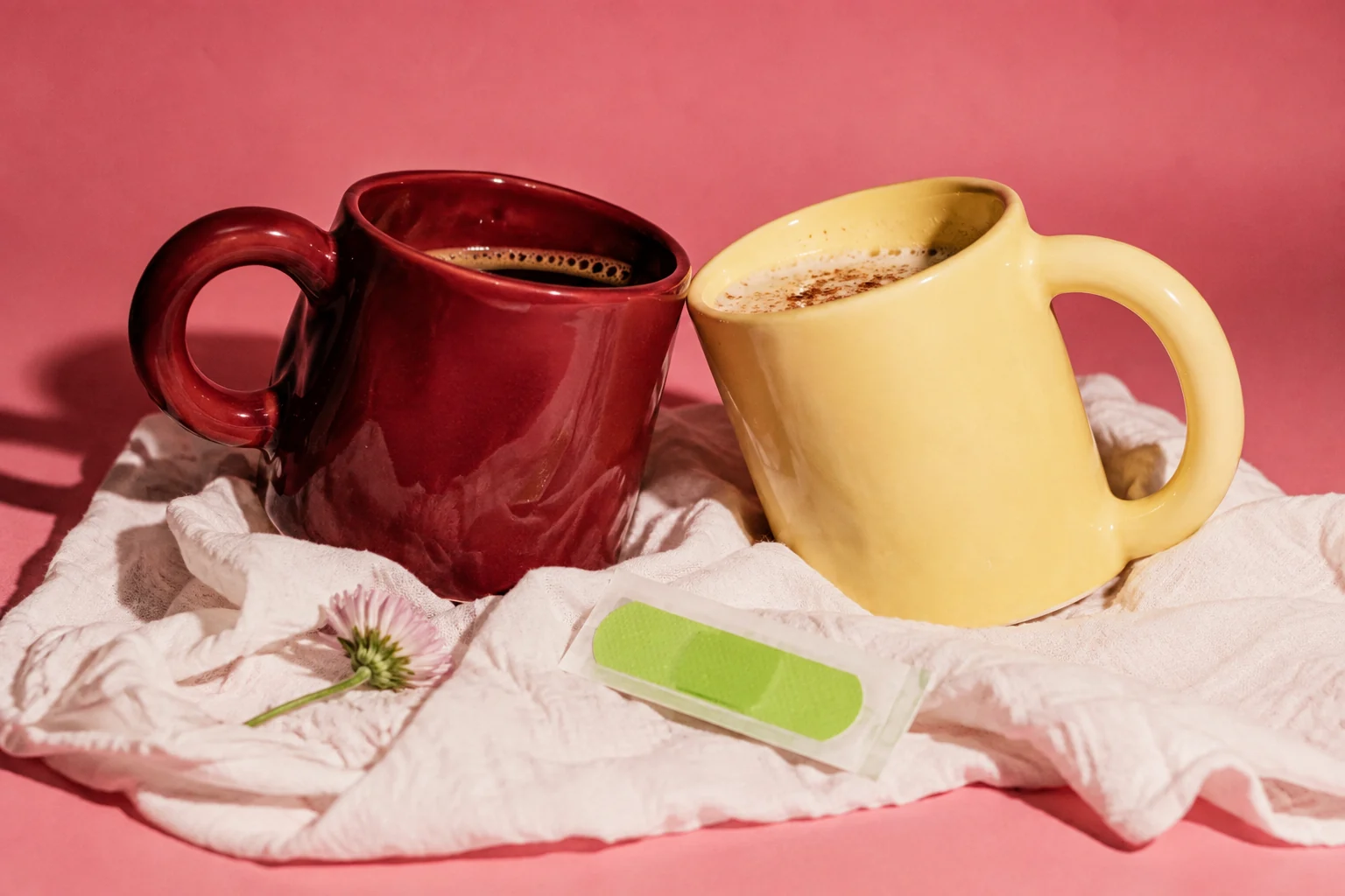 Dos tazas de colores y una tirita sobre tela blanca y fondo rosa.